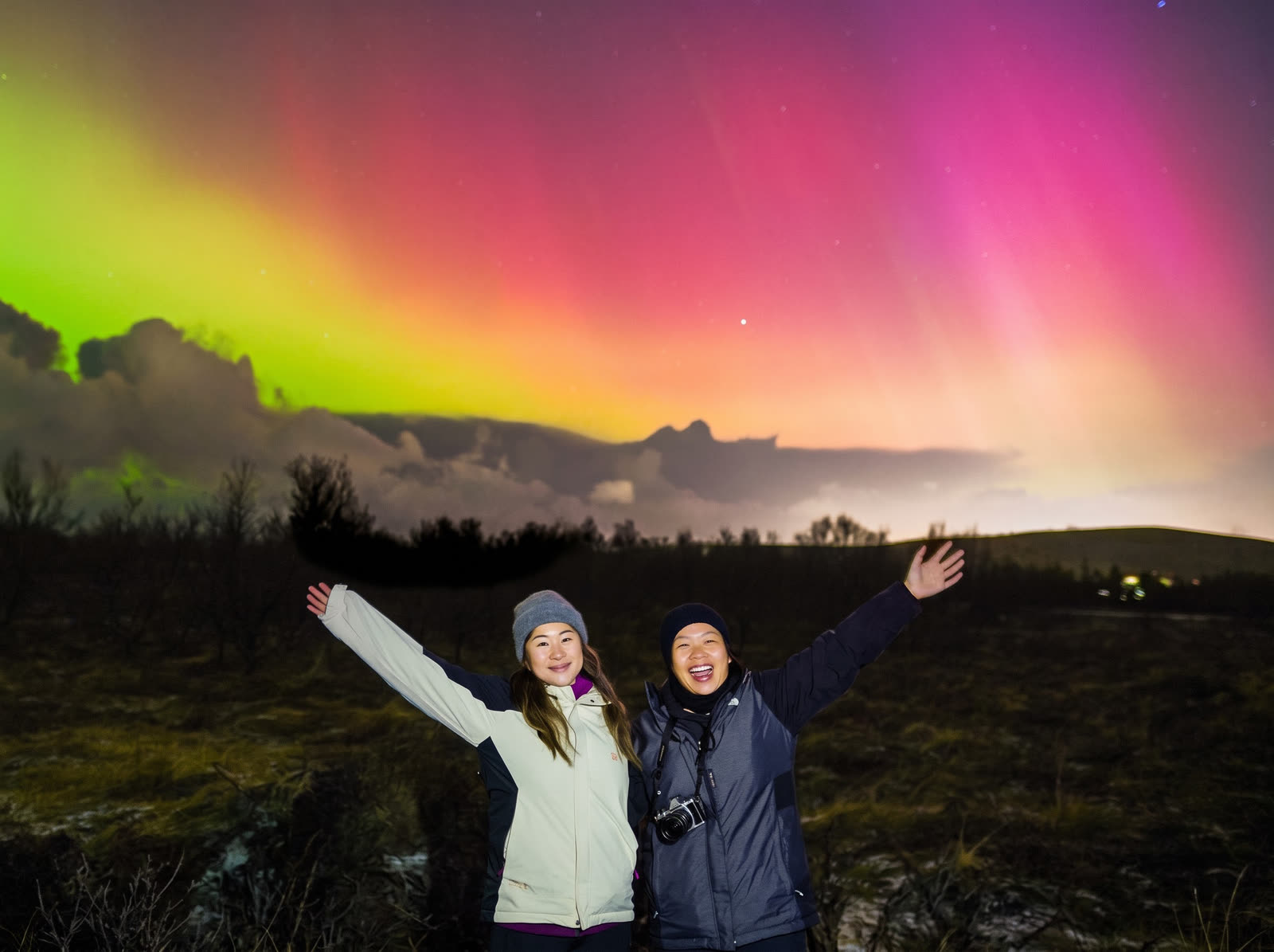 Hai du khách giơ tay chào dưới cực quang hồng và xanh, Iceland